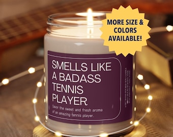Puede incluir: Una vela de vidrio marrón con una etiqueta morada que dice "Smells Like A Badass Tennis Player". La etiqueta también dice "Savor the sweet and fresh aroma of an amazing tennis player." La vela está sentada en un posavasos de madera y está encendida.