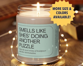 Könnte beinhalten: Eine braune Kerze mit einem weißen Etikett, auf dem "Smells like she's doing another puzzle. Savor the sweet aroma of brainpower and sheer determination." steht. Die Kerze steht auf einem Untersetzer aus Holz. Der Text "More size & colors available!" befindet sich in einem gelben Kreis.