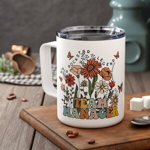 May include: White ceramic mug with a floral design and the text "Team Manager" in colourful retro lettering. The mug has a black handle and a blue lid.