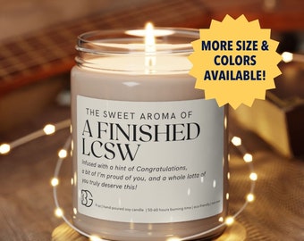 May include: A brown soy candle with a white label that reads "The Sweet Aroma of A Finished LCSW" and "Infused with a hint of Congratulations, a bit of I'm proud of you, and a whole lotta of you truly deserve this!" The candle is lit and sitting on a wooden coaster.