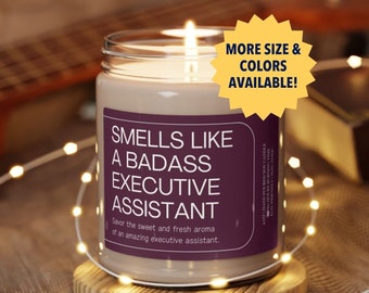 Op de afbeelding: Een bruine glazen kaars met een paars label waarop staat "Smells Like A Badass Executive Assistant". De kaars staat op een houten onderzetter en is omringd door witte lichtsnoeren.