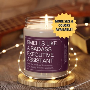 Könnte beinhalten: Eine braune Glaskerze mit einem violetten Etikett, auf dem "Smells Like A Badass Executive Assistant" steht. Die Kerze steht auf einem Holzuntersetzer und ist von weißen Lichterketten umgeben.