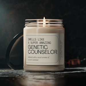 Pode incluir: Uma vela bege num frasco de vidro com uma tampa preta. A vela tem um rótulo branco com o texto "Smells Like A Super Amazing Genetic Counselor" e "Infused with a sweet aroma of a 100% rockstar."
