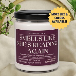 May include: A white soy candle with a black lid and a purple label that reads "Smells Like She's Reading Again". The label also includes the text "Infused with a hint of the sweet rustle of turning pages and the quiet escape of a captivating story."  The candle is hand-poured and eco-friendly. It has a burn time of 50-60 hours.