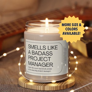 Könnte beinhalten: Eine braune Kerze mit einem weißen Etikett, auf dem "Smells Like A Badass Project Manager. Savor the sweet and fresh aroma of an amazing project manager." steht. Die Kerze steht auf einem Holzuntersetzer und ist von weißen Lichterketten umgeben.
