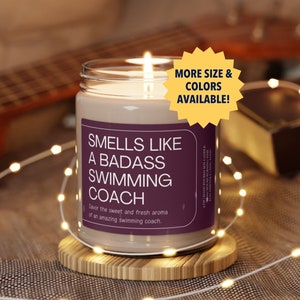 May include: A brown glass candle with a white label that reads "Smells Like A Badass Swimming Coach". The label also says "Savor the sweet and fresh aroma of an amazing swimming coach." The candle is lit and sitting on a wooden coaster. A yellow starburst sticker on the candle says "More Size & Colors Available!"
