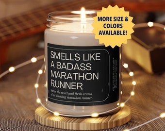 May include: A brown glass candle with a black label that reads "Smells Like A Badass Marathon Runner". The candle is sitting on a wooden coaster and surrounded by white fairy lights.