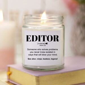May include: A white candle in a clear glass jar with a black label that reads "EDITOR" and a definition of the word. The candle is lit and the flame is visible.