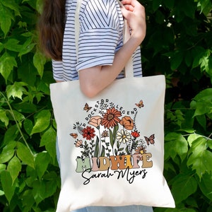 May include: A white canvas tote bag with a floral design and the text "Midwife Sarah Myers". The text is surrounded by flowers and butterflies in shades of orange, pink, yellow, and green. The text "Ninja Rock Star Legend" is written in a circle around the flowers.