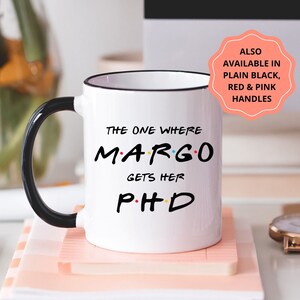 Könnte beinhalten: Eine weiße Keramiktasse mit schwarzem Henkel und schwarzem Rand. Die Tasse trägt den Text "THE ONE WHERE MARGO GETS HER PHD" in schwarzer Schrift. Der Text ist im Stil der Titelkarte der Fernsehserie "Friends" gestaltet.
