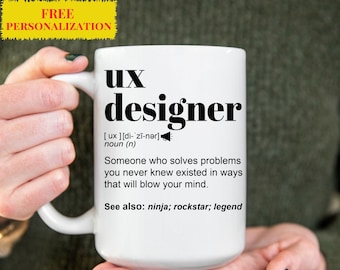 Op de afbeelding: Een witte keramische mok met een zwart ontwerp waarop "UX designer" staat en de term wordt gedefinieerd als "Iemand die problemen oplost waarvan je nooit wist dat ze bestonden, op manieren die je geest zullen doen exploderen." De mok zegt ook "Zie ook: ninja; rockstar; legende".