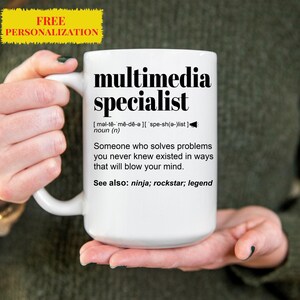 May include: White ceramic coffee mug with a black design that reads "multimedia specialist" and defines the term as "Someone who solves problems you never knew existed in ways that will blow your mind." The mug also includes the text "See also: ninja; rockstar; legend".