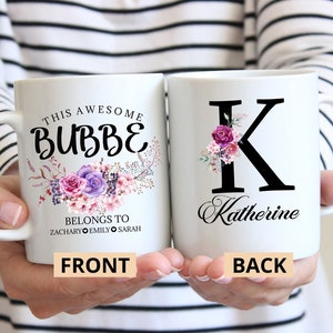May include: Two white ceramic mugs. One mug has the text "THIS AWESOME BUBBE" with floral accents and the names Zachary, Emily, and Sarah. The other mug has a black letter "K" with floral accents and the name Katherine.
