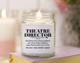 Peut inclure: Une bougie blanche dans un bocal en verre avec une étiquette noire qui indique "THEATRE DIRECTOR" et une définition du mot. La bougie est posée sur une pile de livres.