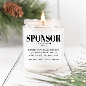 May include: A lit, white candle in a clear glass jar. The label reads "SPONSOR" with a definition. The candle is on a white wooden surface, with greenery and a pinecone in the background.