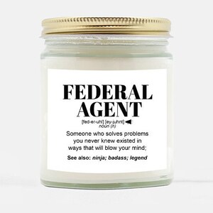 May include: A white candle in a glass jar with a gold lid. The label reads "FEDERAL AGENT [fed-er-uhl] [ey-juhnt] noun (n) Someone who solves problems you never knew existed in ways that will blow your mind; See also: ninja; badass; legend"