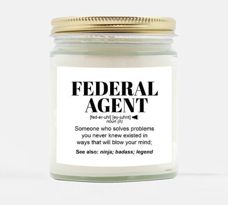 May include: A white candle in a glass jar with a gold lid. The label reads "FEDERAL AGENT [fed-er-uhl] [ey-juhnt] noun (n) Someone who solves problems you never knew existed in ways that will blow your mind; See also: ninja; badass; legend"