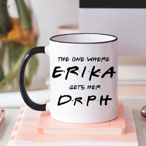 May include: White ceramic mug with a black handle and black rim. The mug has a black graphic that says "The One Where Erika Gets Her DR.PH" with colorful dots around the letters.