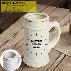 May include: White ceramic beer stein mug with gold-colored detailing. The mug has a handle and a decorative design. The text "CUSTOM DESIGN HERE" is displayed on the front. The mug is 22oz.