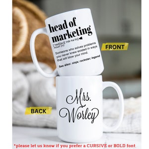 May include: Two white ceramic mugs stacked on top of each other. The top mug reads "head of marketing" with a definition. The bottom mug says "Mrs. Worley" in cursive. The front and back of the mugs are labeled.