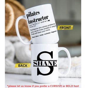 May include: White ceramic mug with a black design. The front of the mug says "pilates instructor" with a definition. The back of the mug has a black letter "S" with the name "SHANE" inside the letter.