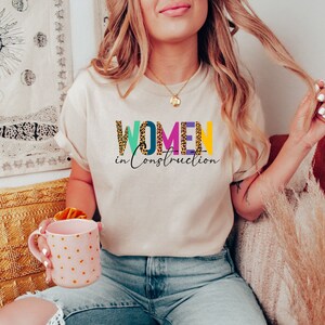 May include: A beige t-shirt with the text "WOMEN in Construction" in colorful letters with a leopard print pattern.