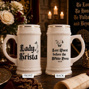 May include: Two white ceramic beer steins with gold rim accents and textured beaded bands. The left stein features the text Lady Krista with a butterfly graphic. The right stein displays the text Last Quest before the White Dress 11.15.26.