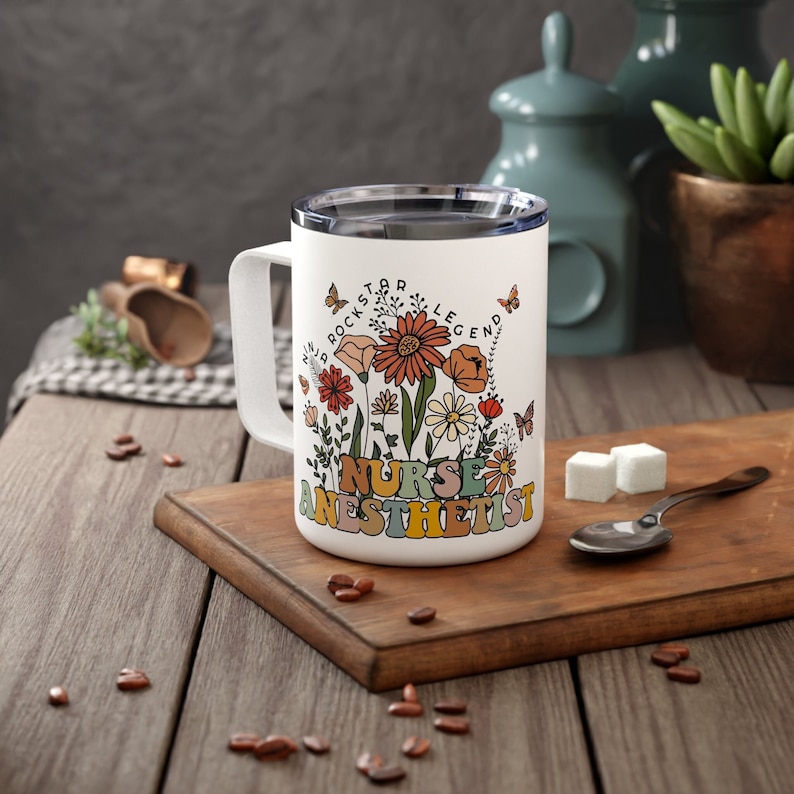 Puede incluir: Taza de viaje de cer&aacute;mica blanca con un dise&ntilde;o floral y el texto "Nurse Anesthetist" en letras de colores. El texto "Rockstar Legend" tambi&eacute;n est&aacute; incluido en el dise&ntilde;o.