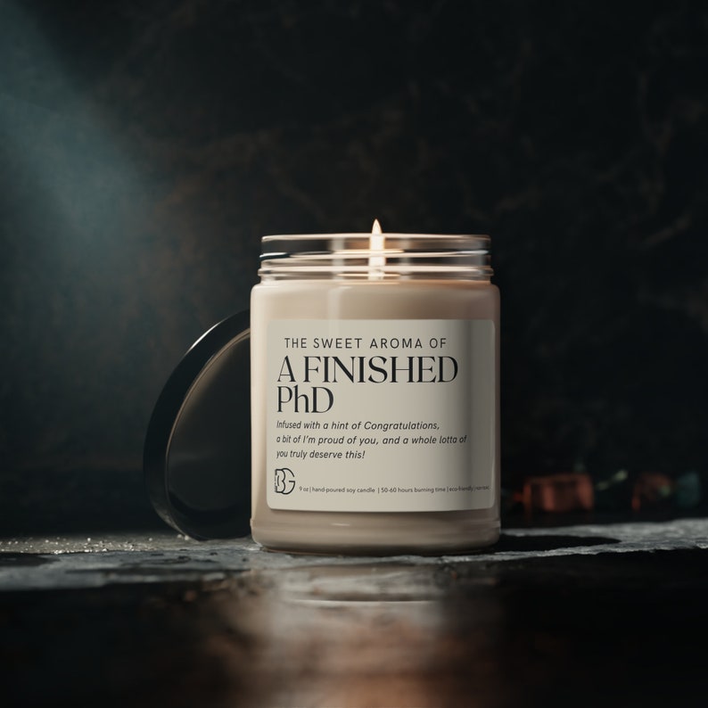 May include: A beige soy candle with a black label that reads "The Sweet Aroma of A Finished PhD" and "Infused with a hint of Congratulations, a bit of I'm proud of you, and a whole lotta of you truly deserve this!"