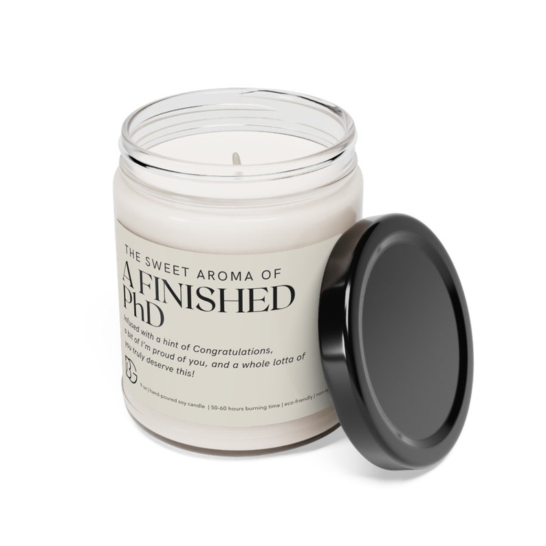 May include: A white soy candle in a clear glass jar with a black lid. The label reads "The Sweet Aroma of A Finished PhD" with a message about congratulations and a hint of pride.