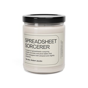 May include: A white soy candle in a glass jar with a black lid. The label reads "SPREADSHEET SORCERER" and describes the candle as "A wizard of spreadsheets conjuring complex formulas and pivot tables that leave colleagues both amazed and slightly perplexed." The label also says "See also: Robert Jacobs".