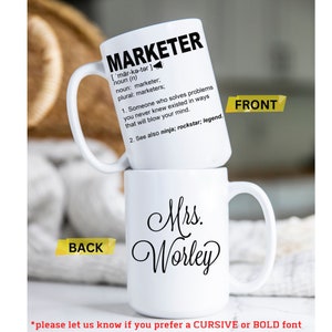 May include: White ceramic coffee mug with a black design. The front of the mug has the word "MARKETER" defined as "Someone who solves problems you never knew existed in ways that will blow your mind." The back of the mug has the text "Mrs. Worley" in cursive.