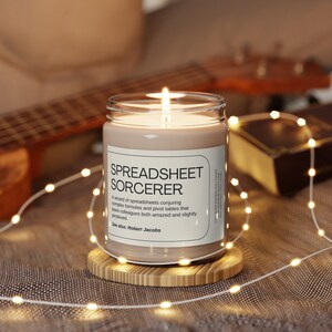 May include: A brown candle with a white label that reads "SPREADSHEET SORCERER" and "A wizard of spreadsheets conjuring complex formulas and pivot tables that leave colleagues both amazed and slightly perplexed. See also: Robert Jacobs".