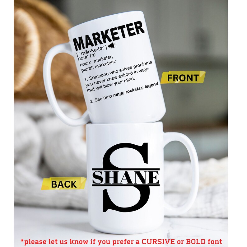 May include: White ceramic coffee mug with a black handle. The front of the mug has the word "MARKETER" written in black with a definition below. The back of the mug has a black letter "S" with the name "SHANE" written across the middle of the letter.