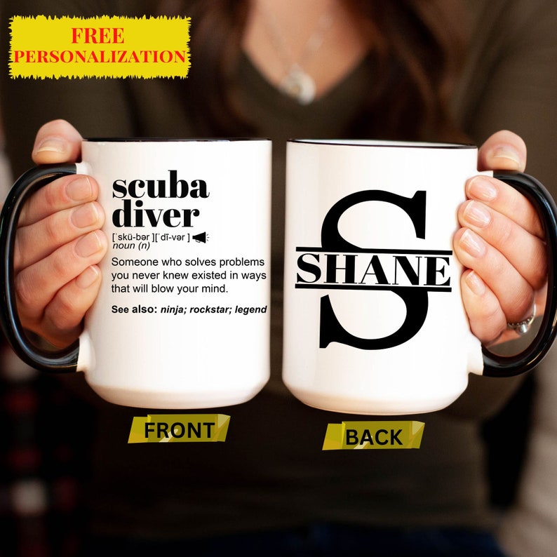 Puede incluir: Taza de cer&aacute;mica blanca con asa y borde negros. La parte delantera de la taza tiene el texto "scuba diver" con una definici&oacute;n y el texto "See also: ninja; rockstar; legend". La parte posterior de la taza tiene una gran letra negra "S" con el nombre "SHANE" escrito en el medio.