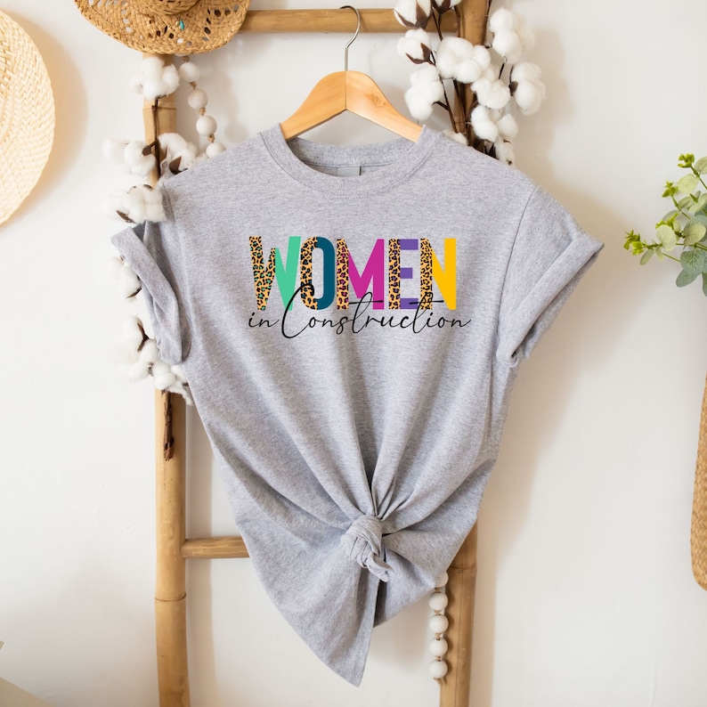 May include: A gray t-shirt with the text "WOMEN in Construction" printed on it. The letters are in different colors and have a leopard print pattern.