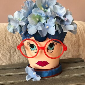 May include: A painted terracotta planter shaped like a face with blue and white flowers on top. The planter has red glasses, blue eyelashes, and a pink mouth.