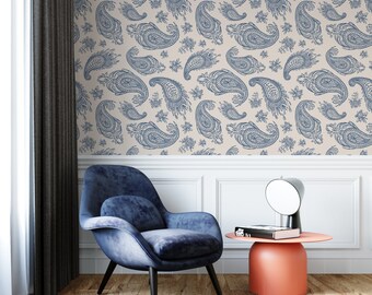 May include: A blue velvet armchair with black legs sits on a grey rug in front of a wall with a beige paisley wallpaper pattern. A small, round, orange side table with a black lamp and a book sits next to the chair.