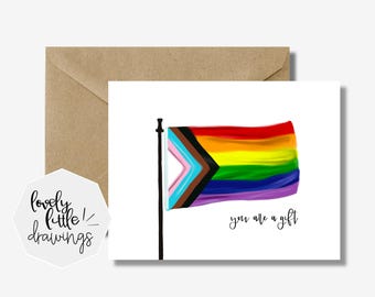 May include: A greeting card with a rainbow flag design, including the Progress Pride flag. The card has the text "you are a gift" in a handwritten font. A brown envelope is behind the card. The card is from "lovely little drawings".