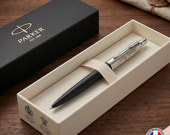 May include: A matte black and silver ballpoint pen with the name Tony Grace engraved on the barrel. The pen rests in a cream-colored presentation box with a matching ribbon, next to a black Parker gift box featuring gold logo text.