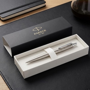 May include: A silver-toned ballpoint pen with the name Caroline Arnold engraved on the barrel, resting in a white tray inside a black Parker gift box featuring the gold Parker Est. 1888 logo on the lid, presented in a rectangular orientation.