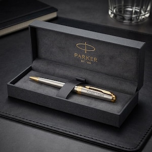May include: A silver and gold ballpoint pen with the name Adam Grant engraved on the barrel, resting inside a dark gray presentation box. The interior lid of the box features the gold Parker logo and the text EST. 1888 in a clean, professional layout.