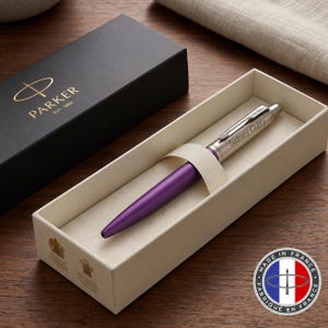 May include: A Parker ballpoint pen with a matte purple barrel and a silver cap engraved with the name Sidney Curry. The pen rests in a cream-colored presentation box next to its black lid, which features the gold Parker logo and text.