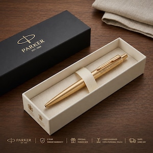 May include: A gold-toned Parker ballpoint pen with the name Bobby Lawrence engraved on the barrel, resting in a cream-colored presentation box. Beside it sits a black Parker gift box featuring the brand logo and the text Parker Est. 1888.