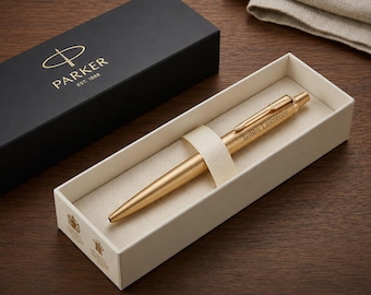 Op de afbeelding: Een goudkleurige Parker-balpen met de naam Bobby Lawrence gegraveerd op de houder, rustend in een crèmekleurige geschenkdoos. Daarnaast staat een zwarte Parker-geschenkdoos met het merklogo en de tekst Parker Est. 1888.
