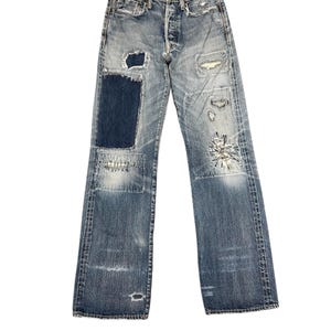 May include: Faded blue denim jeans with multiple patches and distressed details. The jeans have a button fly and a relaxed fit. Patches are visible on the knees and thighs. The jeans have a vintage, worn look.