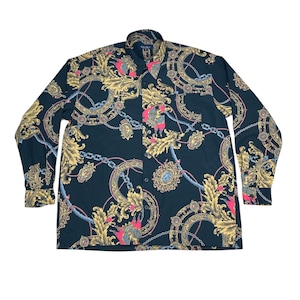 May include: A long-sleeved shirt with a dark background featuring a repeating pattern of gold, pink, and blue ornate designs. The pattern includes chains, medallions, and floral elements. The shirt has a classic collar and buttoned cuffs.