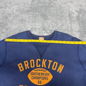 May include: A navy blue sweatshirt with the word "BROCKTON" in orange block letters. Below the word is an orange football graphic with the text "SOUTHERN.DIV CHAMPIONS 68". The word "FOOTBALL" is printed in orange below the graphic.