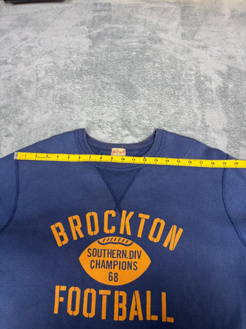 May include: A navy blue sweatshirt with the word "BROCKTON" in orange block letters. Below the word is an orange football graphic with the text "SOUTHERN.DIV CHAMPIONS 68". The word "FOOTBALL" is printed in orange below the graphic.