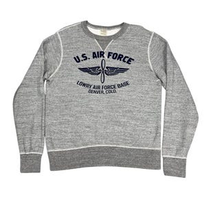 Può includere: Felpa grigia girocollo con la scritta "U.S. AIR FORCE" sopra una grafica di elica e ali. Sotto la grafica c'è la scritta "LOWRY AIR FORCE BASE DENVER, COLO." La felpa ha cuciture bianche e orlo e polsini a coste grigie.